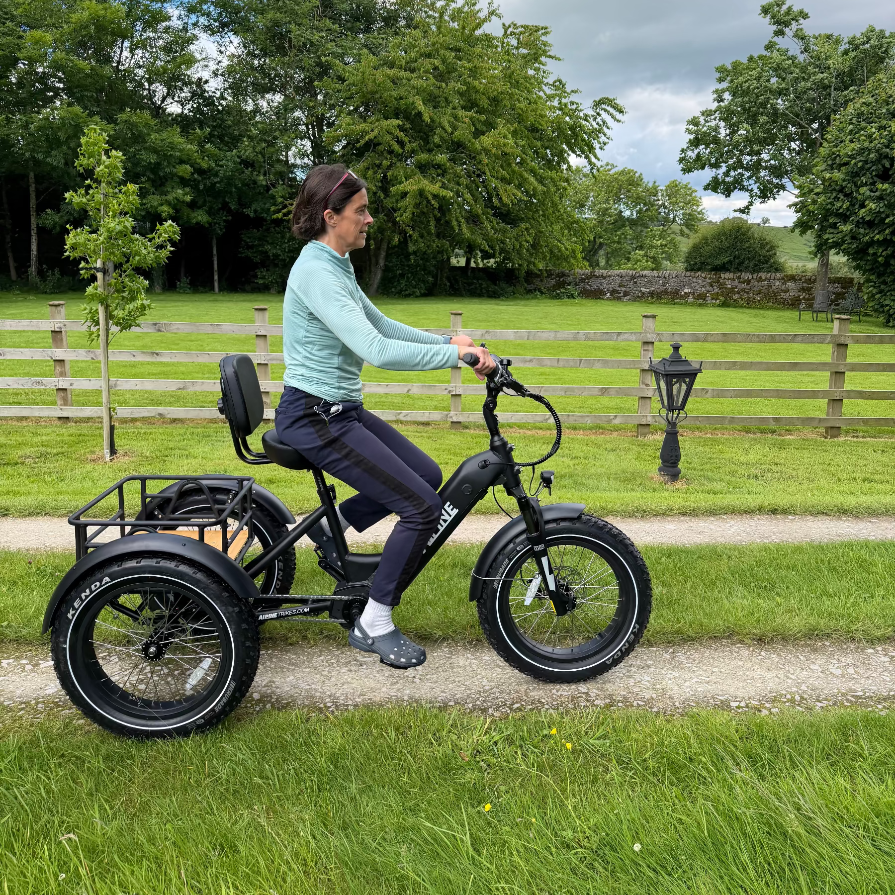 Person riding a black electric tricycle on a path with greenery and a wooden fence in the background.