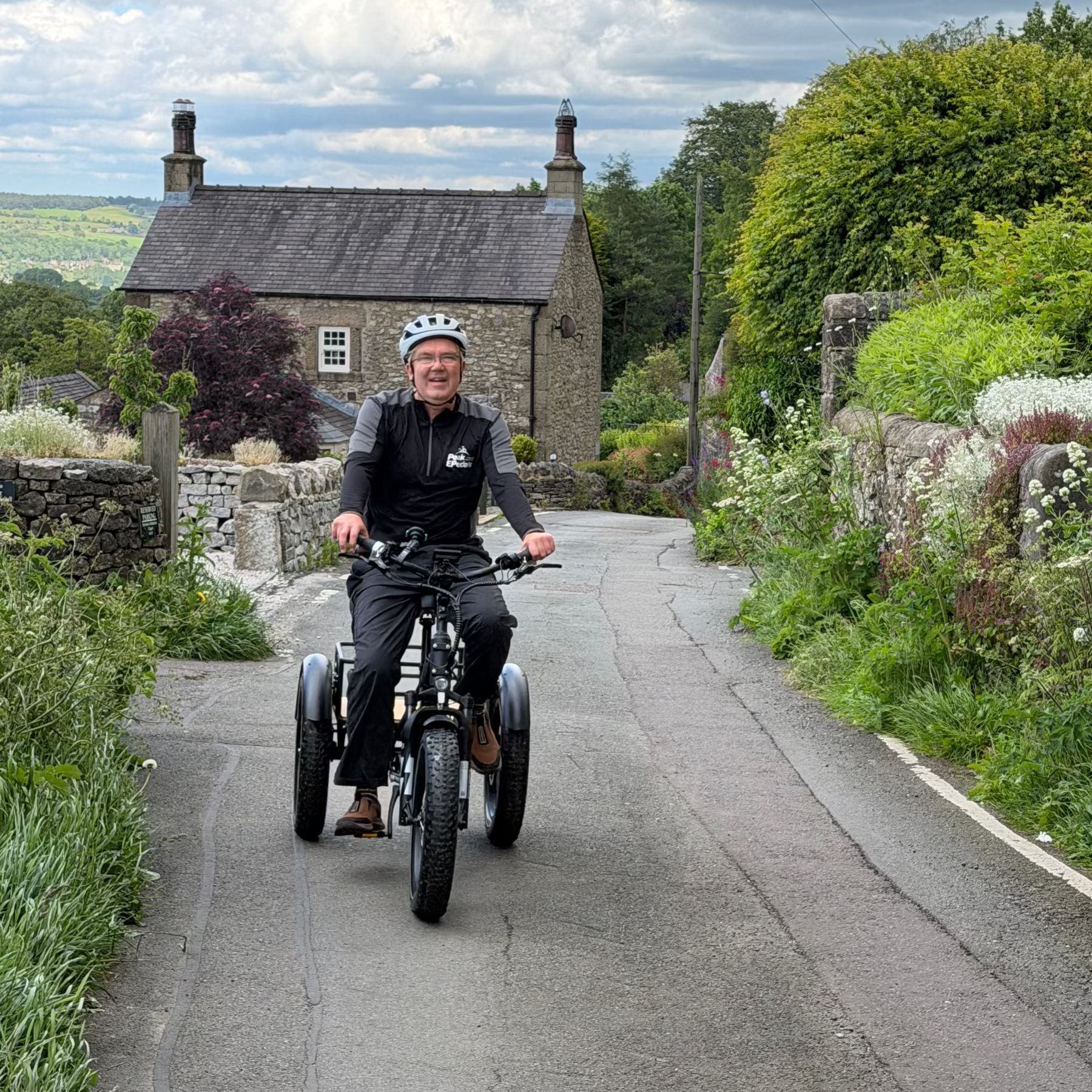 Man riding a tricycle on a rural road with a stone building and greenery in the background.