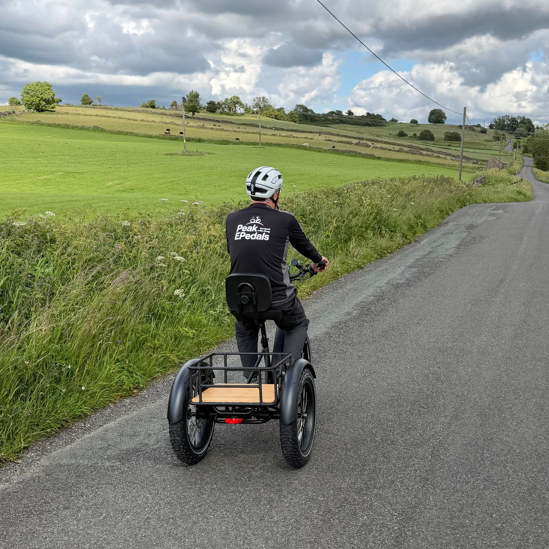 Person riding a three-wheeled trike on a rural road with green fields and cloudy sky.